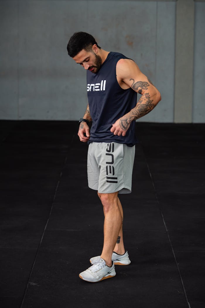 Muscular man in sportswear looking down, showcasing tattoos in an indoor gym setting.
